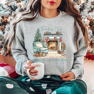 K&ouml;nnte beinhalten: Ein graues Sweatshirt mit einem weihnachtlichen Design, das einen Kamin, einen Weihnachtsbaum und den Text "Readers Club - Cheer and Snuggles Near" zeigt.