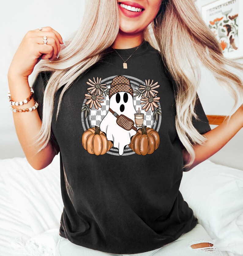 May include: A black t-shirt with a graphic of a ghost wearing a brown and white checkered hat, a brown purse, and holding a brown and white cup. The ghost is surrounded by brown and white flowers and a checkered pattern.