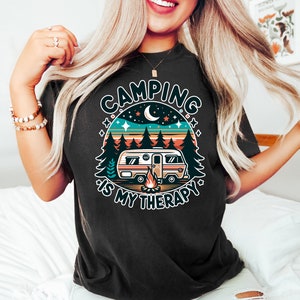 May include: A black t-shirt with a retro-style graphic of a camper van in a forest setting. The graphic includes a campfire, stars, and a crescent moon. The text "Camping is my therapy" is written around the graphic.