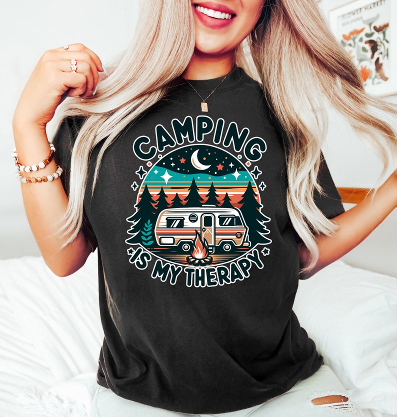 May include: A black t-shirt with a retro-style graphic of a camper van in a forest setting. The graphic includes a campfire, stars, and a crescent moon. The text "Camping is my therapy" is written around the graphic.