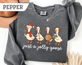 May include: A dark gray sweatshirt with a whimsical illustration of four geese dressed for the holidays. The geese are wearing a Santa hat, Christmas lights, a sweater, and earmuffs. The text "just a jolly goose" is written in cursive below the geese.