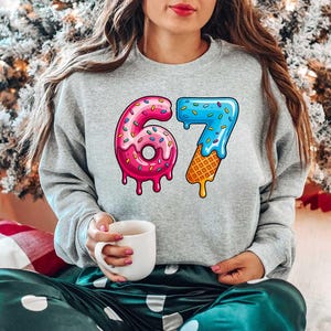 May include: A gray sweatshirt with a colorful graphic of the number 67, designed to look like a donut and ice cream cone. The number 6 is pink with sprinkles, and the 7 is blue with a waffle cone base. The person is holding a white mug.