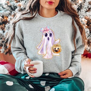 May include: A light gray sweatshirt featuring a cute ghost design holding a jack-o'-lantern. The ghost has a pink bow and gold glitter accents. The sweatshirt is part of a Halloween-themed collection.