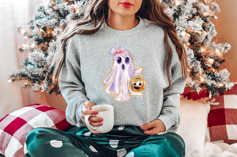 May include: A light gray sweatshirt featuring a cute ghost design holding a jack-o'-lantern. The ghost has a pink bow and gold glitter accents. The sweatshirt is part of a Halloween-themed collection.
