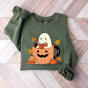 May include: Green sweatshirt featuring a cute ghost reading a book inside a pumpkin mug. The mug is surrounded by autumn leaves and a small pumpkin.