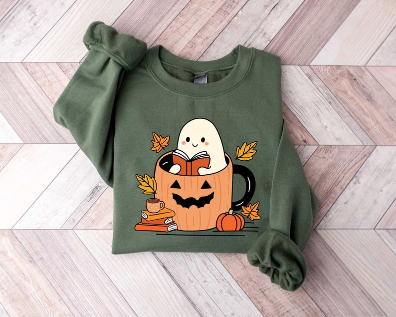 May include: Green sweatshirt featuring a cute ghost reading a book inside a pumpkin mug. The mug is surrounded by autumn leaves and a small pumpkin.