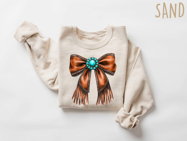 May include: A beige sweatshirt with a brown leather bow with turquoise gemstone embellishments and fringe details. The word "SAND" is printed in brown text in the upper right corner of the image.