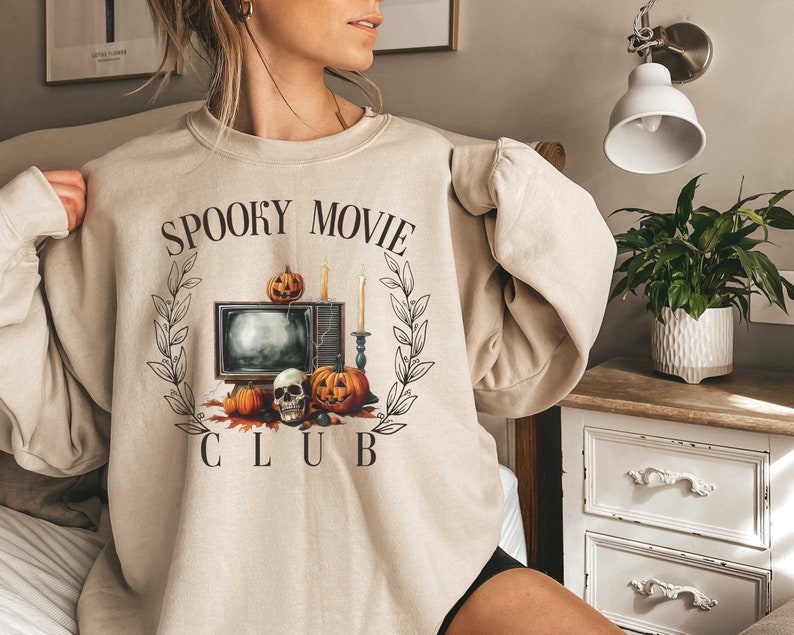 K&ouml;nnte beinhalten: Ein beiger Pullover mit einem grafischen Aufdruck eines Fernsehers, K&uuml;rbissen und einem Sch&auml;del. Der Text "Spooky Movie Club" ist auf dem Pullover aufgedruckt.