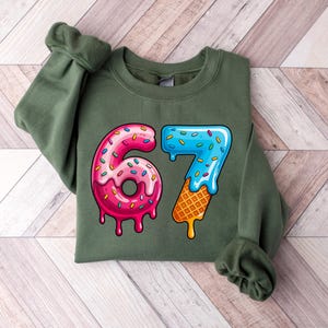 May include: An olive green sweatshirt with a colorful graphic of the numbers 67. The number 6 is designed as a pink donut with sprinkles, and the number 7 is designed as a blue ice cream cone with a waffle cone base.