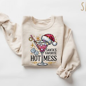 May include: A beige sweatshirt with a festive design featuring a martini glass with a red drink, ice cubes, and a Santa hat. The text "Santa's Favorite Hot Mess" is printed below the glass.