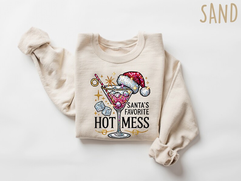 May include: A beige sweatshirt with a festive design featuring a martini glass with a red drink, ice cubes, and a Santa hat. The text "Santa's Favorite Hot Mess" is printed below the glass.