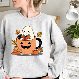 May include: A grey sweatshirt featuring a cute ghost reading a book inside a pumpkin mug. The ghost is smiling and holding a book. The mug is surrounded by autumn leaves and a small pumpkin.
