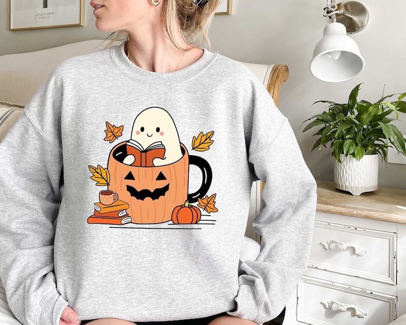 May include: A grey sweatshirt featuring a cute ghost reading a book inside a pumpkin mug. The ghost is smiling and holding a book. The mug is surrounded by autumn leaves and a small pumpkin.