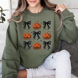 Op de afbeelding: Een groen sweatshirt met een Halloween-thema met zwarte strikken en oranje pompoenen met jack-o'-lantern gezichten.