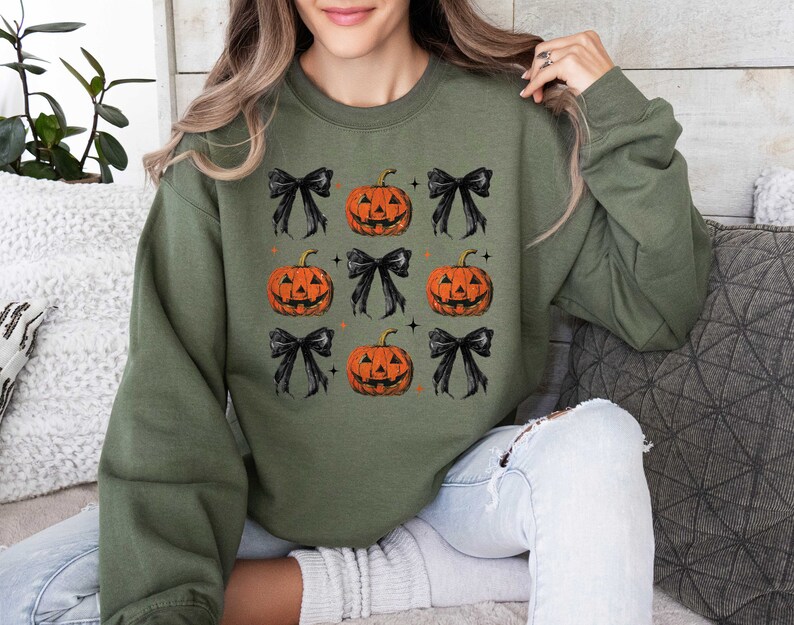 Op de afbeelding: Een groen sweatshirt met een Halloween-thema met zwarte strikken en oranje pompoenen met jack-o'-lantern gezichten.