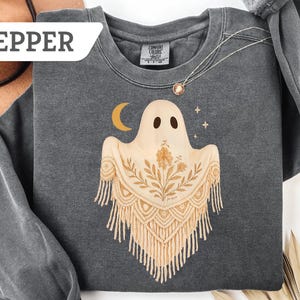 May include: Dark gray sweatshirt with a ghost graphic. The ghost has a beige shawl with floral and fringe details, a crescent moon, and stars. The word "PEPPER" is in a white rectangle in the upper left corner. A gold necklace is visible.