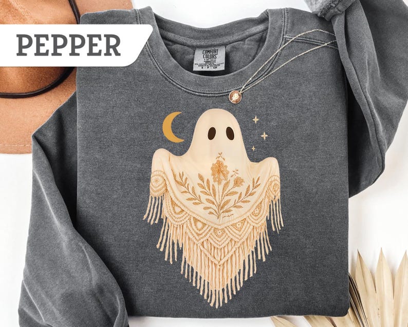 May include: Dark gray sweatshirt with a ghost graphic. The ghost has a beige shawl with floral and fringe details, a crescent moon, and stars. The word "PEPPER" is in a white rectangle in the upper left corner. A gold necklace is visible.