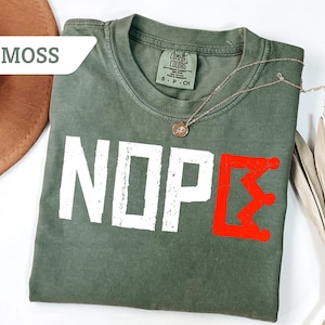 May include: A moss green t-shirt with the word "NOPE" in white block letters and a red graphic. A gold necklace with a pendant rests on the shirt. The shirt is folded and placed on a white surface.