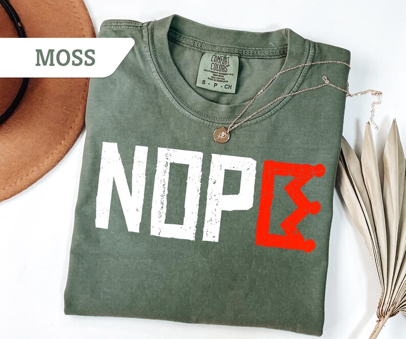 May include: A moss green t-shirt with the word "NOPE" in white block letters and a red graphic. A gold necklace with a pendant rests on the shirt. The shirt is folded and placed on a white surface.