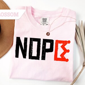 May include: A light pink t-shirt with the word "NOPE" printed in black, and a red crown-like design. A gold necklace with a pendant rests on the shirt. A brown hat and a dried palm leaf are also in the image.