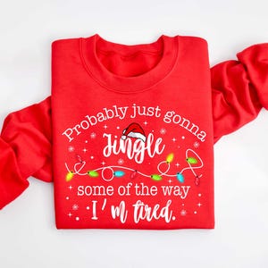 Peut inclure: Sweat-shirt rouge avec le texte "Probably just gonna jingle some of the way, I'm tired." Le motif comprend un bonnet de P&egrave;re No&euml;l, des guirlandes lumineuses et des flocons de neige. V&ecirc;tement de f&ecirc;te sur le th&egrave;me des f&ecirc;tes.