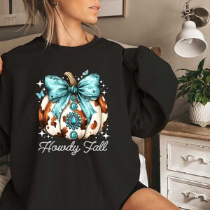Puede incluir: Sudadera negra con una calabaza de estampado de vaca con un lazo azul y pedrer&iacute;a. El texto "Howdy Fall" est&aacute; debajo de la calabaza.