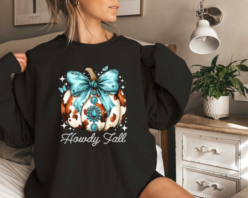 Puede incluir: Sudadera negra con una calabaza de estampado de vaca con un lazo azul y pedrer&iacute;a. El texto "Howdy Fall" est&aacute; debajo de la calabaza.