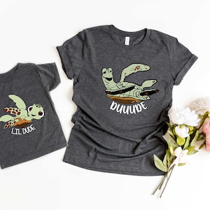 May include: Two grey t-shirts featuring a cartoon sea turtle design. The larger shirt says "DUDE" and the smaller shirt says "LIL DUDE".