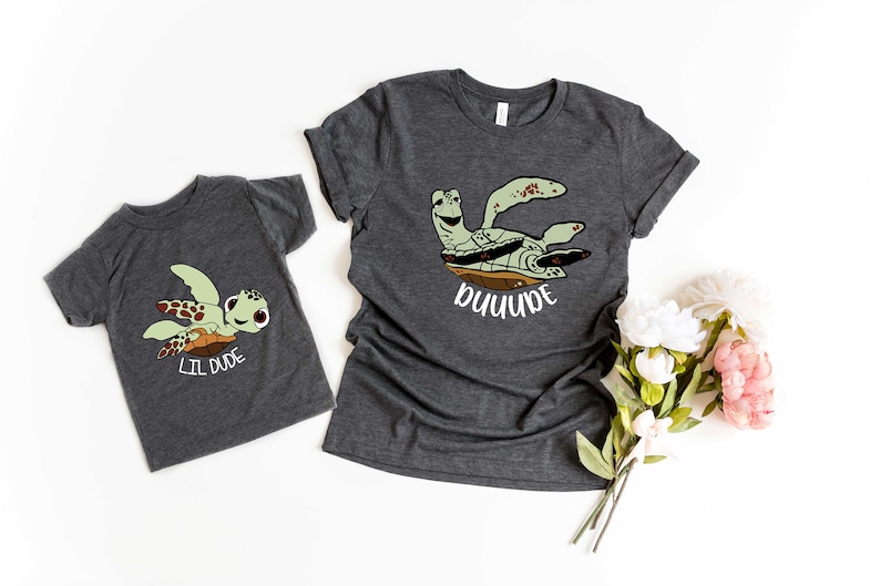 May include: Two grey t-shirts featuring a cartoon sea turtle design. The larger shirt says "DUDE" and the smaller shirt says "LIL DUDE".