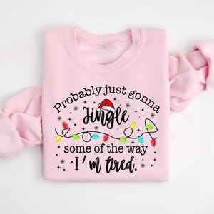 Peut inclure: Un sweat-shirt rose clair avec l'inscription "Probably just gonna jingle some of the way. I'm tired." Le mot "Jingle" est surmont&eacute; d'un bonnet de P&egrave;re No&euml;l, et des guirlandes lumineuses color&eacute;es sont en dessous.