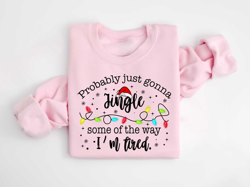 Peut inclure: Un sweat-shirt rose clair avec l'inscription "Probably just gonna jingle some of the way. I'm tired." Le mot "Jingle" est surmont&eacute; d'un bonnet de P&egrave;re No&euml;l, et des guirlandes lumineuses color&eacute;es sont en dessous.