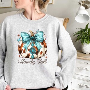 Puede incluir: Una sudadera gris con una calabaza de estampado de vaca marr&oacute;n y blanco con un lazo turquesa y pedrer&iacute;a. El texto "Howdy Fall" est&aacute; impreso debajo de la calabaza.