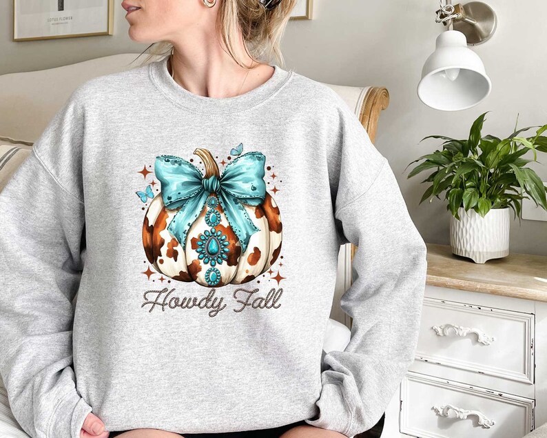 Puede incluir: Una sudadera gris con una calabaza de estampado de vaca marr&oacute;n y blanco con un lazo turquesa y pedrer&iacute;a. El texto "Howdy Fall" est&aacute; impreso debajo de la calabaza.