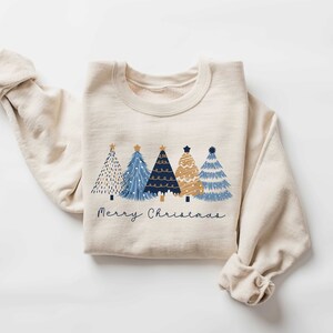 May include: A beige sweatshirt with a graphic of four blue and gold Christmas trees and the text "Merry Christmas".