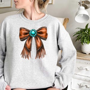 May include: A gray sweatshirt with a brown leather bow with turquoise gemstone embellishment and fringe detail.