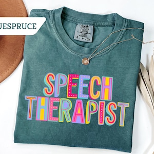 May include: A teal Comfort Colors t-shirt with the words "SPEECH THERAPIST" in colorful, block letters. The shirt has a crew neck and short sleeves. A gold necklace with a pendant is draped across the shirt. A brown hat is visible in the background.
