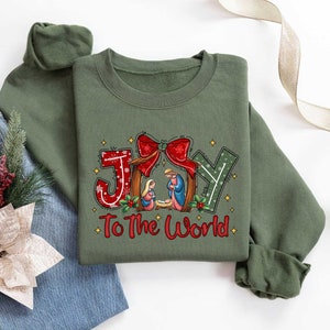 May include: A green sweatshirt with a Christmas-themed design featuring the words "Joy to the World" and a depiction of the nativity scene. The design includes a red bow, a star, and a manger with Mary, Joseph, and baby Jesus.