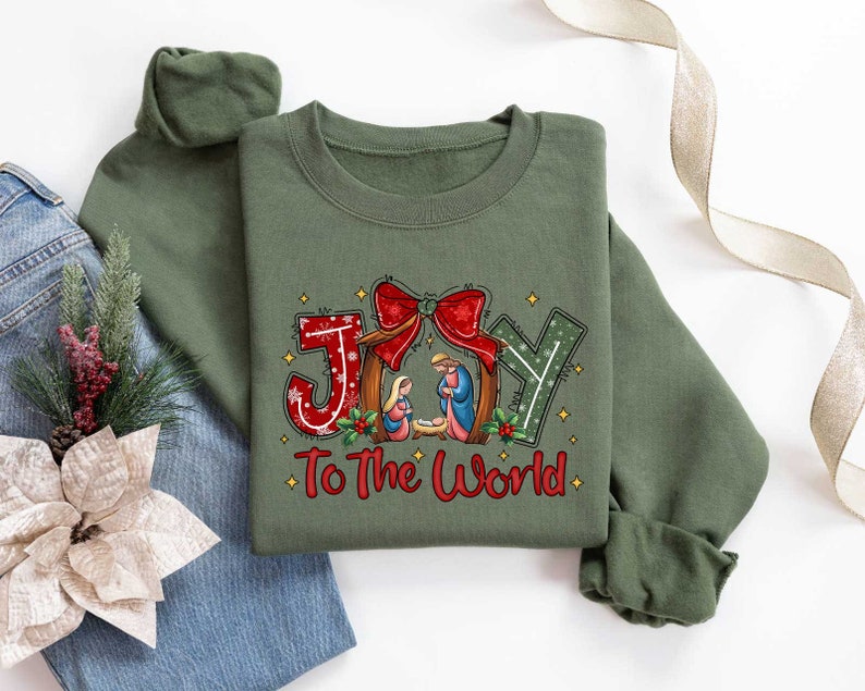 May include: A green sweatshirt with a Christmas-themed design featuring the words "Joy to the World" and a depiction of the nativity scene. The design includes a red bow, a star, and a manger with Mary, Joseph, and baby Jesus.