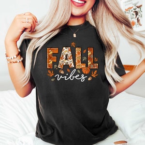 Może przedstawiać: Czarny T-shirt z nadrukiem graficznym słowa "FALL" w kolorze pomarańczowym, brązowym i ż&oacute;łtym z wzorem liści, żołędzi i dyni. Słowo "vibes" jest nadrukowane pod słowem "FALL" na biało.