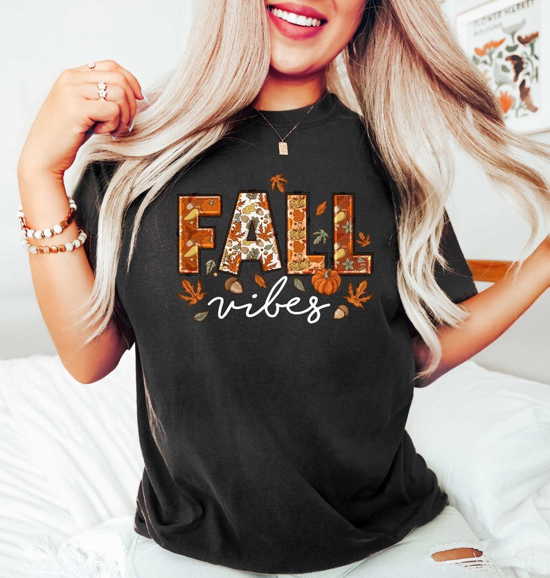 Może przedstawiać: Czarny T-shirt z nadrukiem graficznym słowa "FALL" w kolorze pomarańczowym, brązowym i ż&oacute;łtym z wzorem liści, żołędzi i dyni. Słowo "vibes" jest nadrukowane pod słowem "FALL" na biało.