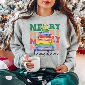 May include: A gray sweatshirt with the words "MERRY teacher" and a colorful Christmas tree design. The tree is made of stacked books with rainbow-colored pages. The sweatshirt is worn by a person holding a white mug.