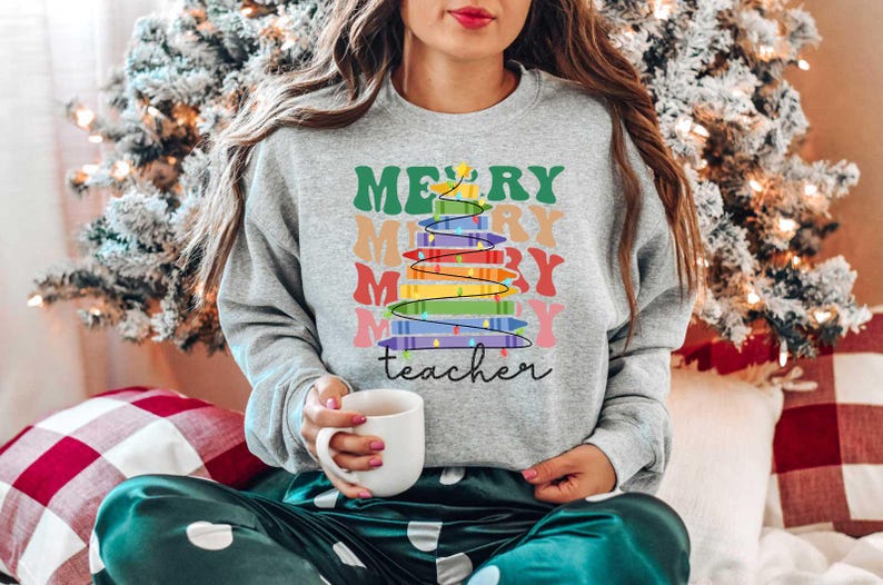 May include: A gray sweatshirt with the words "MERRY teacher" and a colorful Christmas tree design. The tree is made of stacked books with rainbow-colored pages. The sweatshirt is worn by a person holding a white mug.