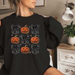 Op de afbeelding: Zwart sweatshirt met een herhalend patroon van oranje pompoenen met zwarte strikken en witte sterren.