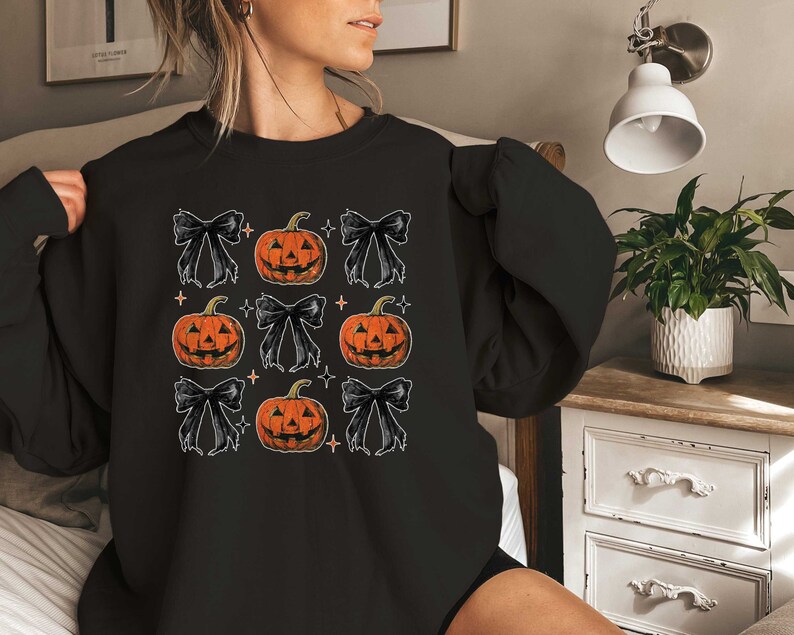 Op de afbeelding: Zwart sweatshirt met een herhalend patroon van oranje pompoenen met zwarte strikken en witte sterren.
