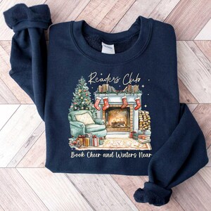 K&ouml;nnte beinhalten: Navyblauer Sweatshirt mit einem weihnachtlichen Design mit einem Kamin, einem Weihnachtsbaum und dem Text "Readers Club Book Cheer and Winters Near".