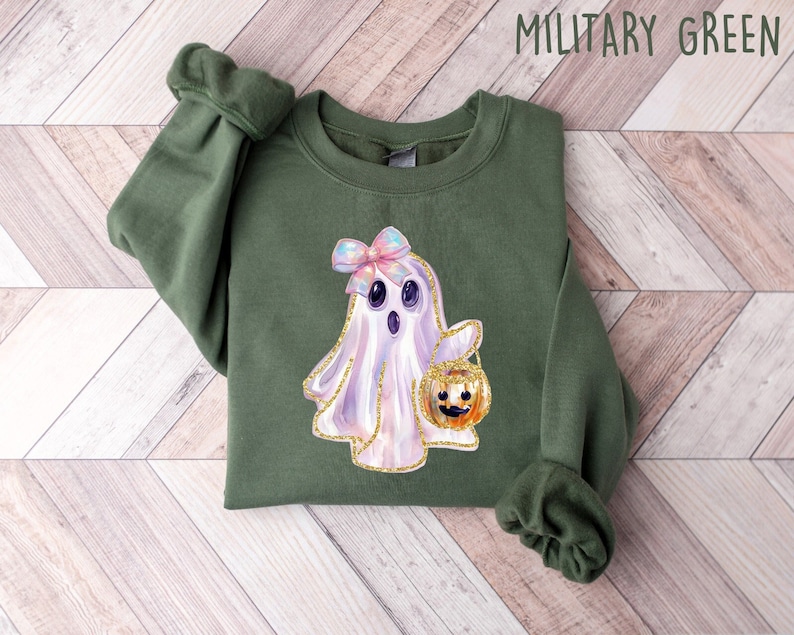 May include: Military green sweatshirt featuring a watercolor ghost with a bow and a pumpkin bucket. The ghost has a gold glitter outline. The text "MILITARY GREEN" is at the top of the image. The sweatshirt is on a light wood background.