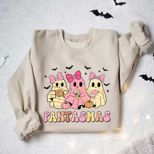 May include: A beige sweatshirt with a graphic of three pink and yellow ghosts holding various treats. The text "PANTASMAS" is printed in pink and yellow letters below the ghosts.