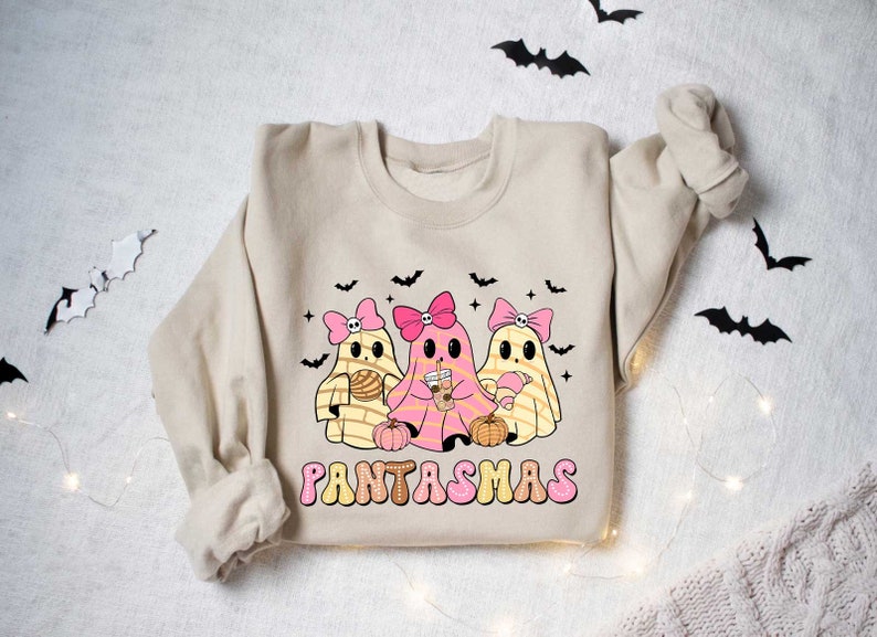 May include: A beige sweatshirt with a graphic of three pink and yellow ghosts holding various treats. The text "PANTASMAS" is printed in pink and yellow letters below the ghosts.