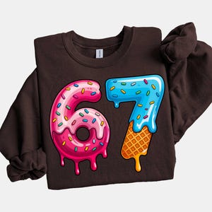 May include: A dark brown sweatshirt featuring a colorful graphic of the number "67". The "6" is designed as a pink donut with sprinkles, and the "7" is blue with a waffle cone base. The design has a dripping effect.