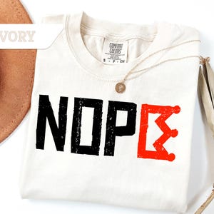 May include: Ivory-colored t-shirt with the word "NOPE" printed in black, and a red crown graphic. The shirt is folded, with a gold necklace and a hat visible in the background. The shirt is made of a soft material.